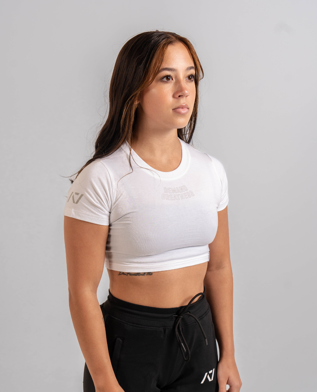 A woman wearing a white crop top with a Demand Greatness design on the upper chest and A7 logo on the upper back and sleeves.