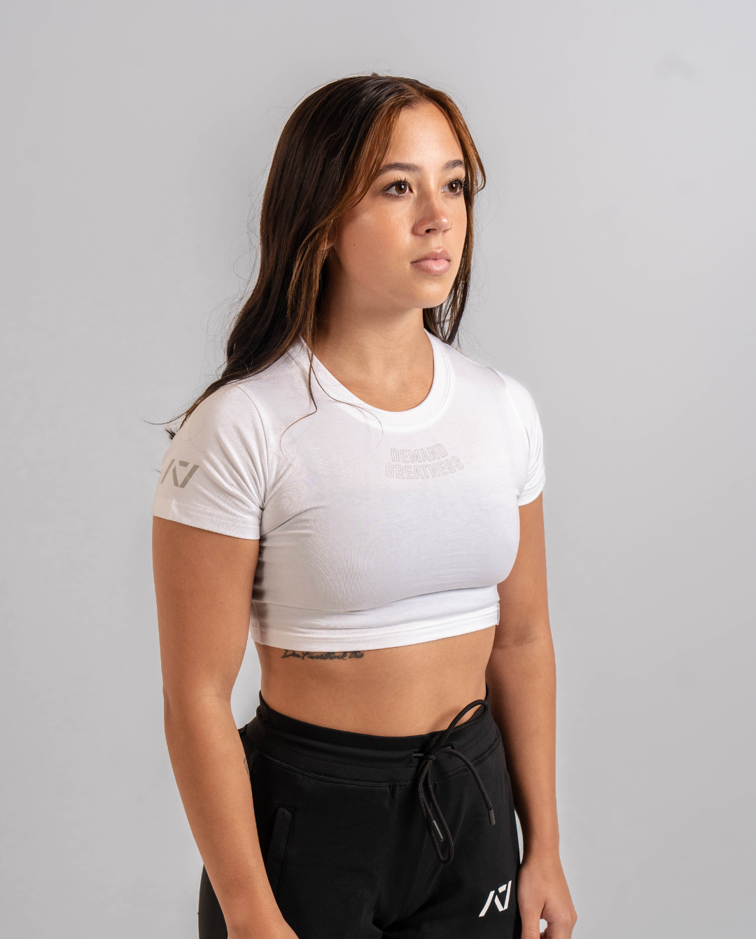 A woman wearing a white crop top with a Demand Greatness design on the upper chest and A7 logo on the upper back and sleeves.