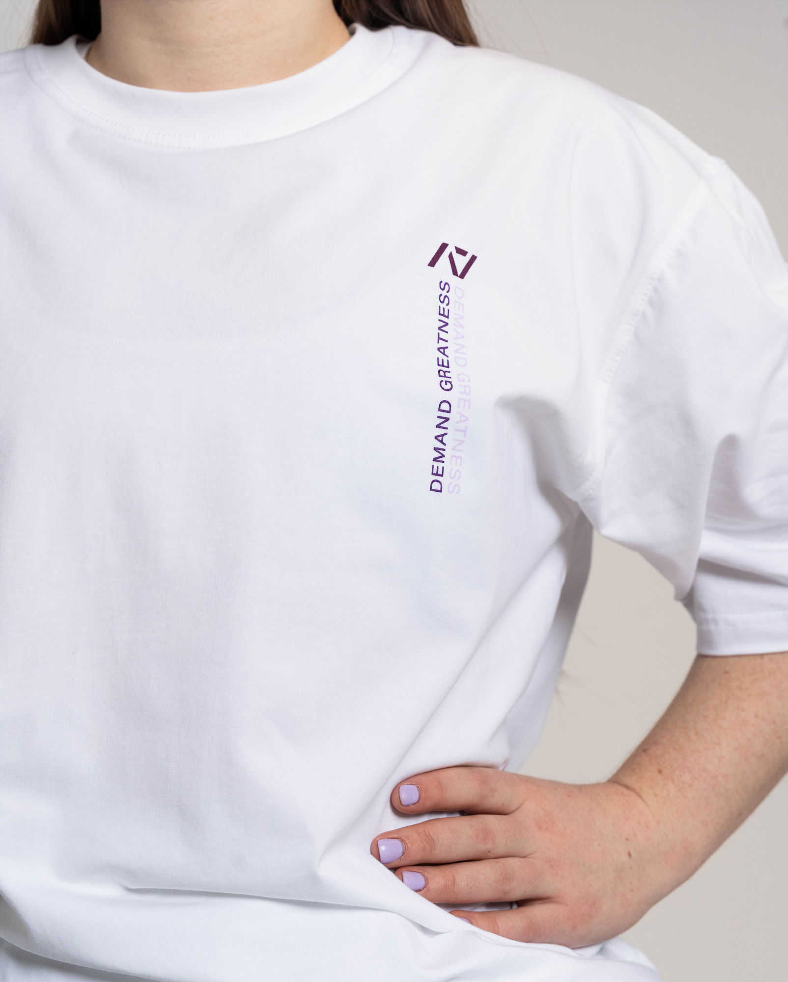 White oversized t-shirt with a logo and text worn by a person, on a neutral background