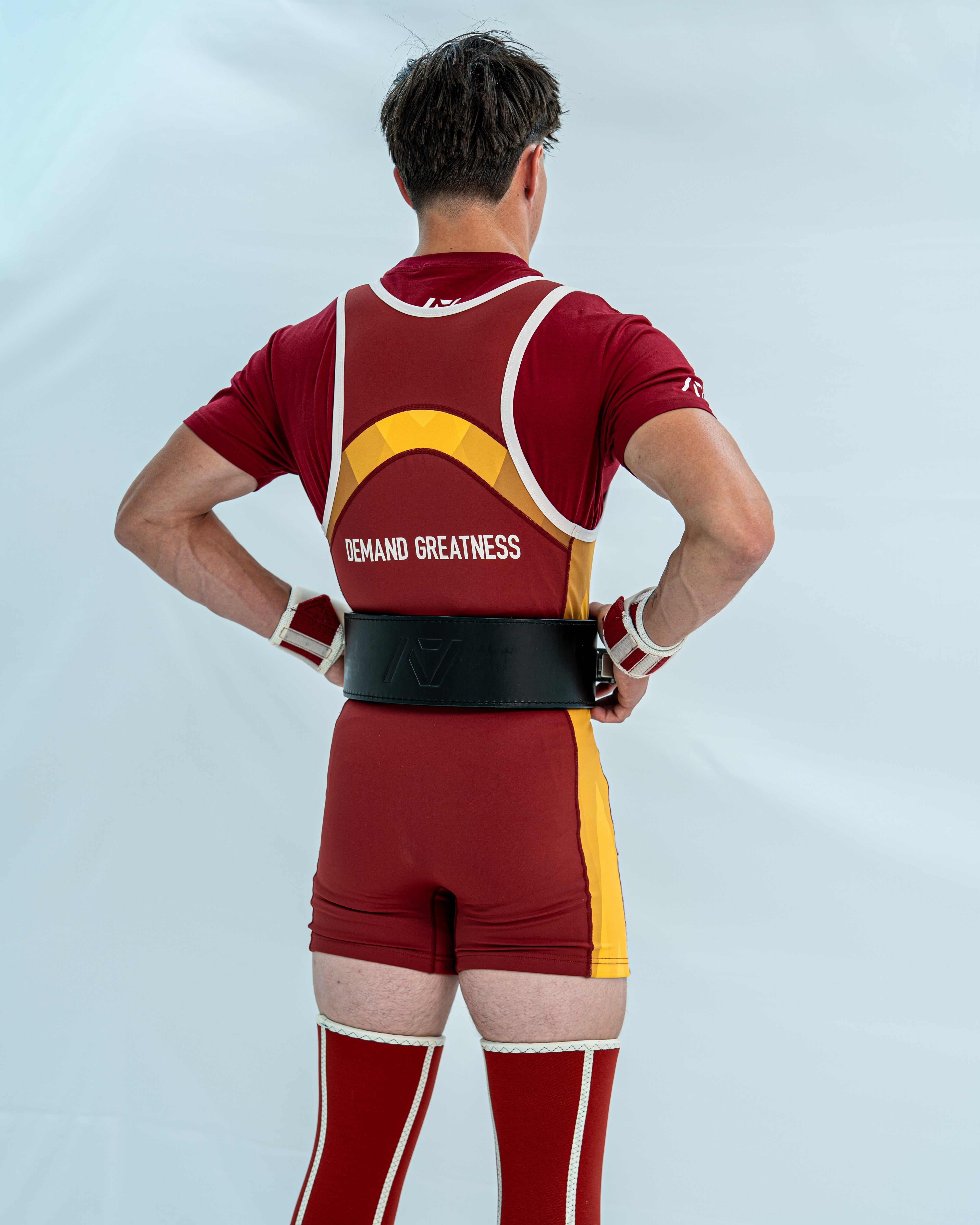 Male model wearing a red and yellow powerlifting singlet with 'Demand Greatness' on the back of the singlet on a light grey background
