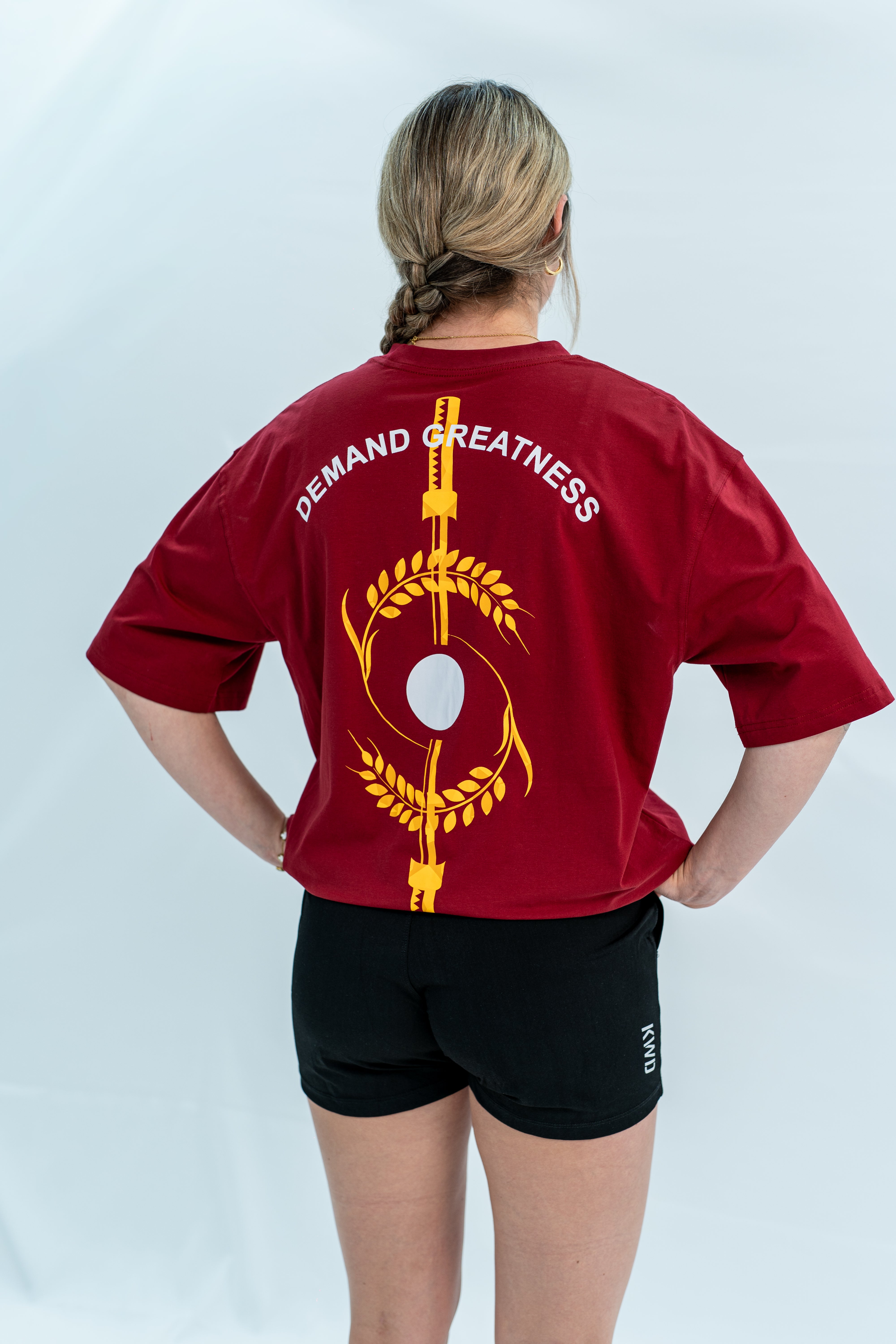 Female wearing a red unisex Over-Fit Shirt with 'Demand Greatness' text and design on the back, standing against a gray background.