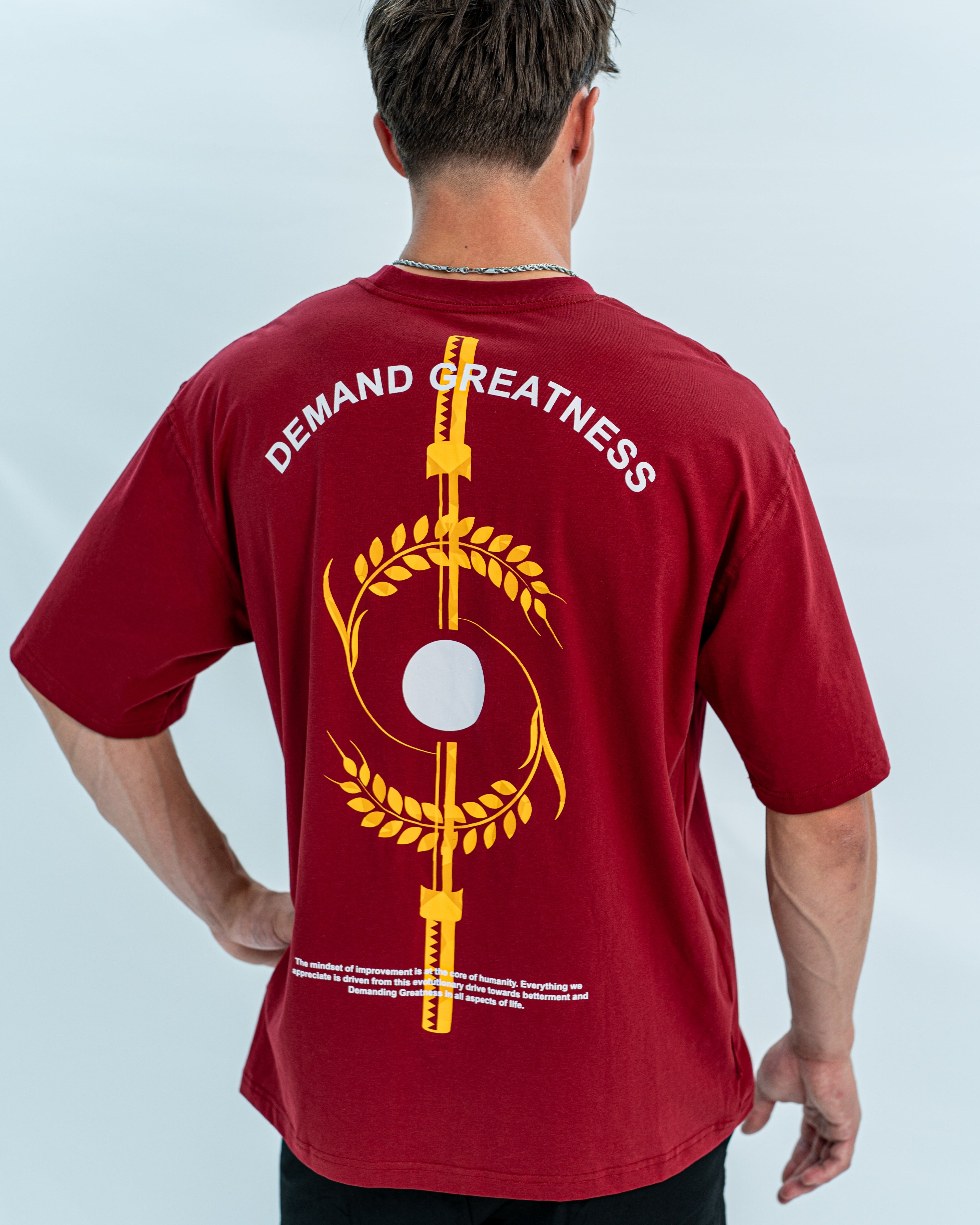 Red over-fit shirt with 'Demand Greatness' text and graphic on the back with equinox design, worn by a person against a light gray background