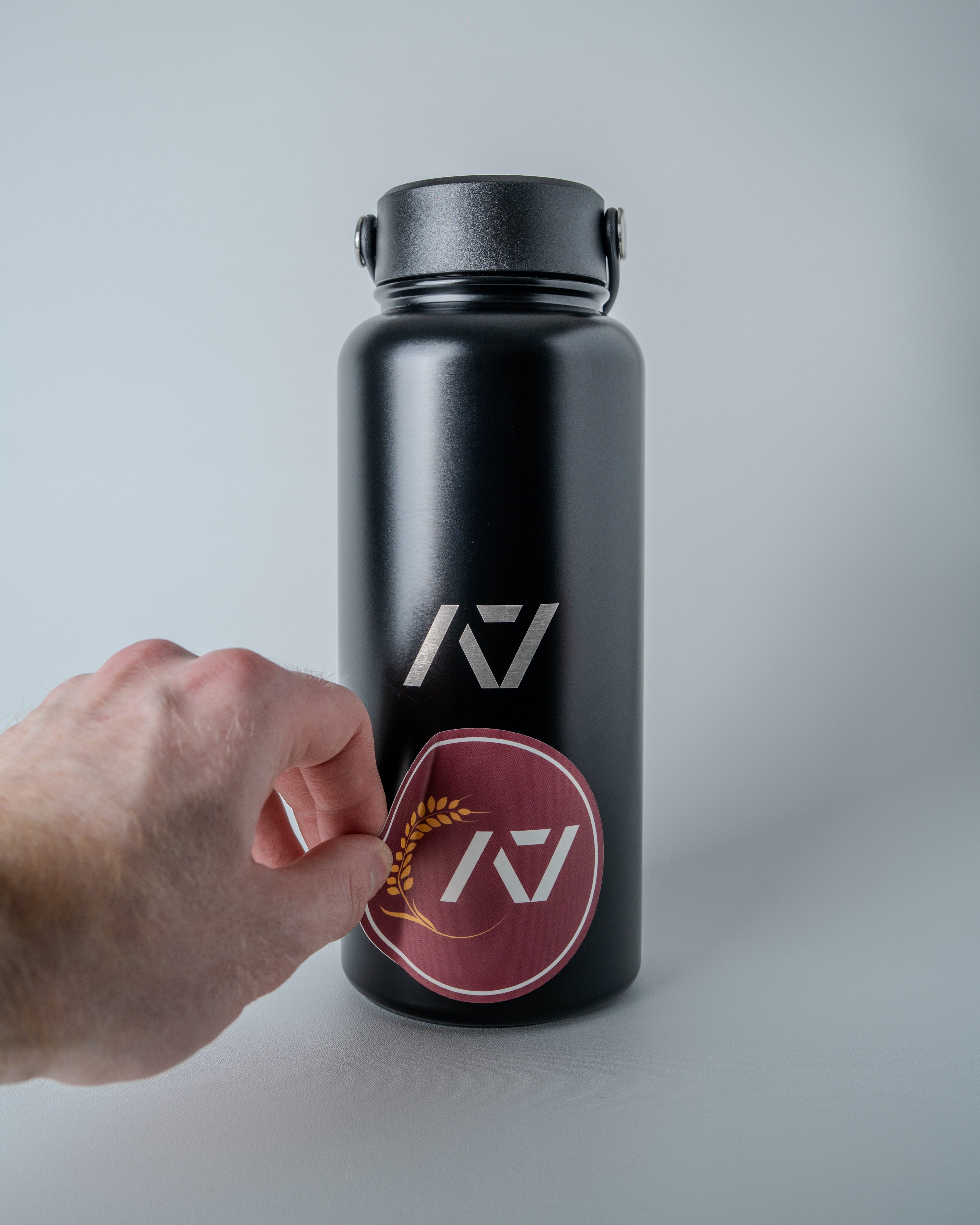 Black water bottle with sticker getting placed onto the bottle on a gray background