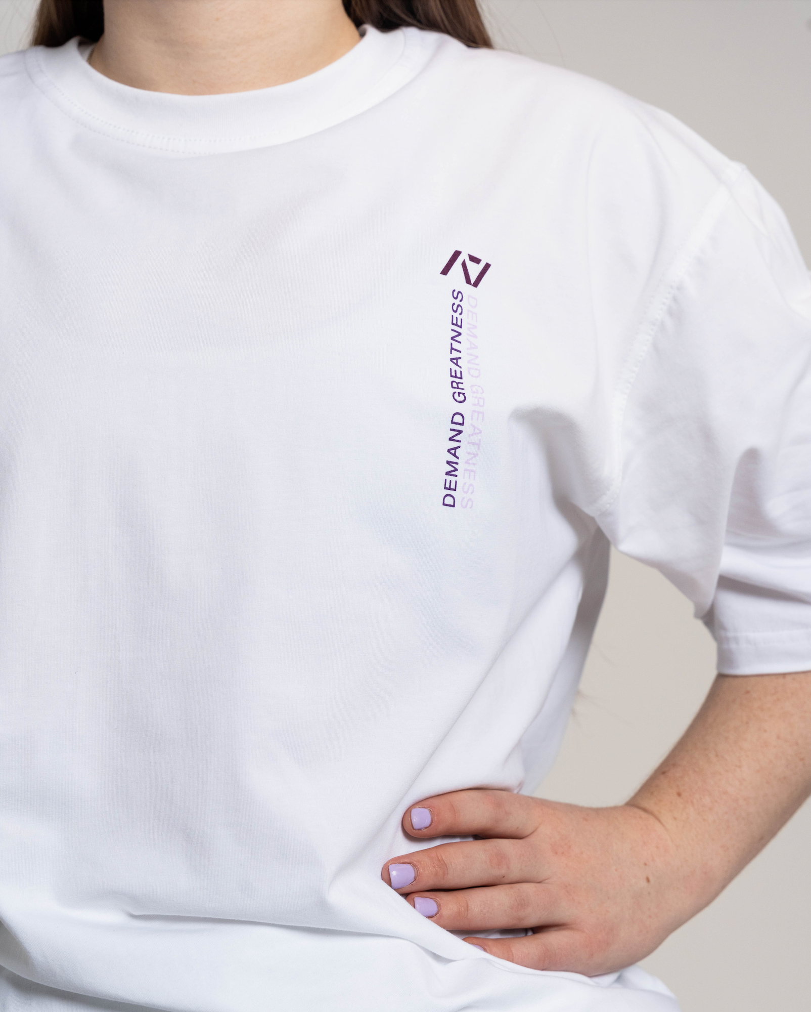 White oversized t-shirt with a logo and text worn by a person, on a neutral background